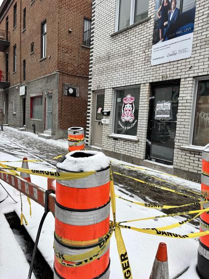 Une piste cyclable enneigée et bloquée par des barrières de chantier et du ruban de signalisation. À l'arrière-plan, on aperçoit des bâtiments en briques et en briques blanches, dont l'un affiche une annonce immobilière.