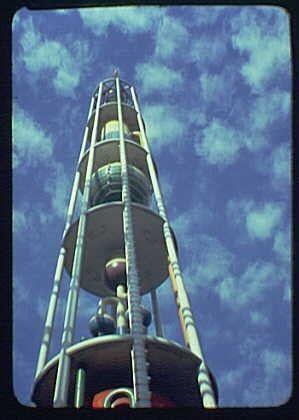 The image depicts a tall, spiraling tower that is reminiscent of a Ferris wheel or observation structure. The coloration suggests it might be an amusement park ride from the mid-20th century, specifically around 1940 based on its design and context provided by additional information in World's Fair texts.
Visually impaired users can imagine this as a towering metallic construction with multiple levels of platforms ascending to form spirals. Each platform appears enclosed or surrounded by glass barriers for safety reasons, typical of an observation tower at the time. The structure is painted primarily in white, possibly indicating its material could be metal, likely steel given historical context.
The sky behind it is blue and dotted with fluffy clouds, suggesting a clear day which enhances visibility from such heights. This imagery can evoke feelings associated with amusement parks or fairgrounds during that era, where innovation and design were showcased through grand structures like the Tower of Chemistry by DuPont Building at an early World's Fair in 1940.
Please note that specific details about the overall size of this structure are not discernible from the given image.