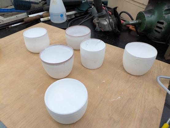 A photo of 6 differently-sized cups sitting on a wooden board. The cups are bright white, except for around the rims where the red/brown terracotta is showing through.