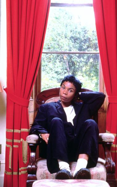 Cedella Marley sitzt vor einem roten Vorhang und einem Fenster auf einem Sessel. Ihre Beine sind hochgelegt. Sie trägt einen schwarzen Hosenanzug.