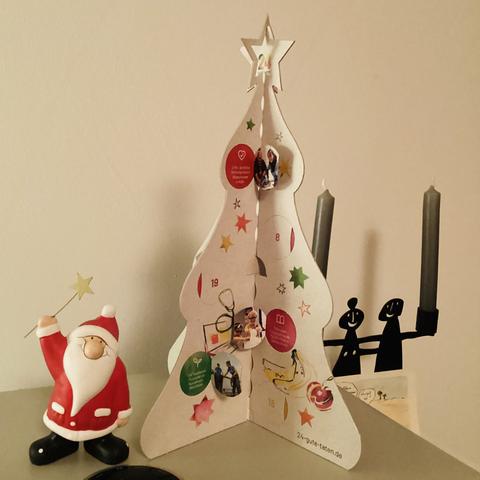 Ein Adventskalender in Form eines Weihnachtsbaums zum Aufstellen. Er ist aus grauem Recycling-Papier gefertigt und bunt bedruckt. Auf dem Weihnachtsbaum sind 24 Türchen angebracht, hinter denen sich die sozialen Projekte verstecken, für die mit dem Kauf gespendet wird. Links davon steht eine kleine Weihnachtsmannfigur. Rechts dahinter ein stilisierter Kerzenständer (Mann + Frau) mit zwei grauen Kerzen. Das  fünfte Türchen des Adventskalenders ist geöffnet.