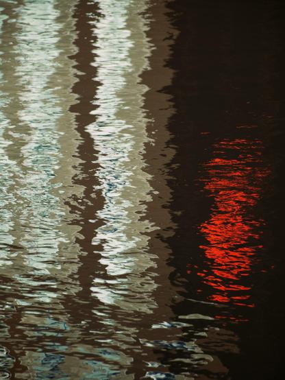 Lights from office buildings and a traffic light are reflected in vertical columns in the shining, rippled surface of a river running through a city centre, looking very much like an abstract painting