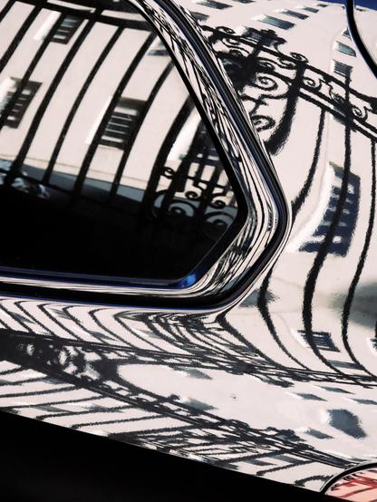 Ornate iron railings and a white building caught in bright sunlight are reflected in the shiny paintwork and window of a parked car in a largely monochrome image with just two tiny hints of colour, blue and red.
