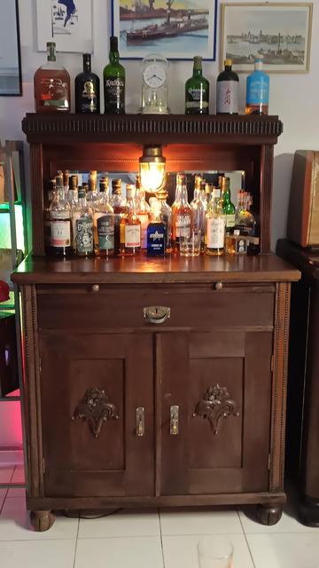 Ein rund 100 Jahre alter Schrank mit Spiegelaufsatz und zentraler Kellerlampe aus Messing vor dem Spiegel. Ebenfall vor dem Spiegel auf der großen Abstellfläche befinden sich zahlreiche Whiskyflaschen, die von der Lampe erleuchtet werden. 
Der Schrank ist in dunklem Holz, wahrscheinlich Nußbaum gehalten.
Die Beschläge sind aus Messing und verfügen jeweils über einen Griff zum einfachen anfassen.
Etwas versteckt sind 2 Knäufe aus Messing, die an eine Ausziehplatte montiert sind, welche aktuell eingeschoben ist.