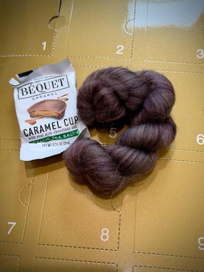 Behind, advent calendar box in gold with numbers in each perforated door. On top, a caramel chocolate cup. To the right, a braid of brownish-purple fiber