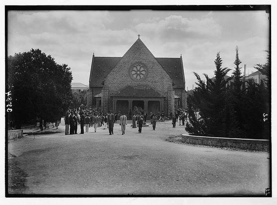 This black and white photograph depicts a group of people exiting a large church-like building with a distinctive pointed roof and arched window above the entrance. The architecture suggests European influence, possibly Gothic or Romanesque in style. Trees flank both sides of the path leading away from the structure, which appears to be situated within an urban environment as buildings are visible on either side.

The crowd is dressed in early 20th-century clothing, with men wearing suits and ties while women wear long dresses, indicating a formal occasion or event such as church services. The individuals appear diverse but all seem orderly as they move from the building towards the camera's perspective. This scene likely represents an American University of Beirut (AUB) chapel service where students are exiting after attending religious ceremony around 1920 to 1933.

The image is labeled with handwritten text, possibly indicating a cataloging or classification system used by its creator or archive for identification and organization purposes during the time it was captured. The photo has some wear on the edges but retains much of its original detail, preserving historical context and visual elements from that era.