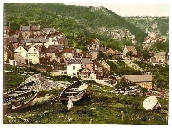 The image depicts a picturesque coastal village nestled in hilly terrain. Traditional stone buildings with steep, gabled roofs are clustered along the slopes of a hillside overlooking an expanse of greenery and scattered rocks or outcrops suggestive of limestone geology typical to this region. In the foreground, two small rowboats sit idle on the grassy shore; one boat is upright while another leans against its hull, both displaying signs of wear and weathering indicative of frequent use. A pile of what appears to be crushed stones sits nearby.

A couple of tents or temporary shelters with open sides are pitched in the vicinity, hinting at possible fishing activity or leisure camping by locals or visitors. The overall atmosphere is serene yet bustling with a sense of lived-in history. It's likely that this image captures Runswick Bay during late 19th to early 20th-century times when such coastal communities were integral parts of local economies centered around maritime activities, possibly including fishing and boatbuilding.

The photograph carries the impression of being part of an album or collection documenting British Isles scenery from a bygone era. The coloration is sepia-toned with subtle hues that provide contrast against the rich greenery and stone architecture, adding to its vintage appeal.