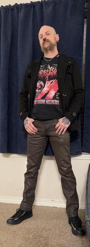 A person with a distinctive appearance, including prominent facial piercings and styled facial hair, is standing in front of a dark blue curtain. They wear a black graphic t-shirt featuring a detailed design that says GhostBath at the top and Rose Thorn Necklace on the bottom with an illustration of lacerated bloody hands and arms in the middle, a black peacoat, and brown denim pants. Their hands have red, black, and blue pawprint tattoos, and they are wearing black sneakers.