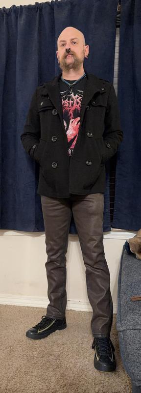 A person stands against a backdrop of dark curtains, wearing a black peacoat over a black graphic t-shirt featuring red artwork. They have a prominent mustache and facial piercings, paired with brown denim pants and black sneakers.