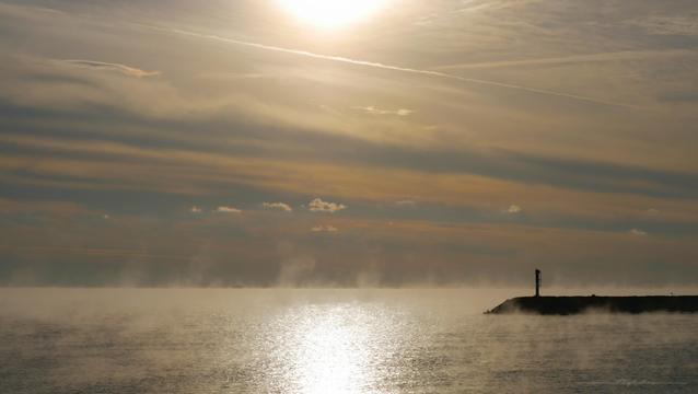 Photo de la mer au petit matin. Sur la droite, on voit une digue à contre jour surmontée d'une balise. Le ciel est barré de différentes couches de nuages aux tons gris et jaunâtres plus ou moins épaisses, et on voit le soleil derrière les nuages en haut au centre de l'image. La mer, qui a pris des reflets argentés, est couverte de fines volutes de vapeur blanche qui s'élèvent vers le ciel et donnent à la photo une ambiance mystérieuse.