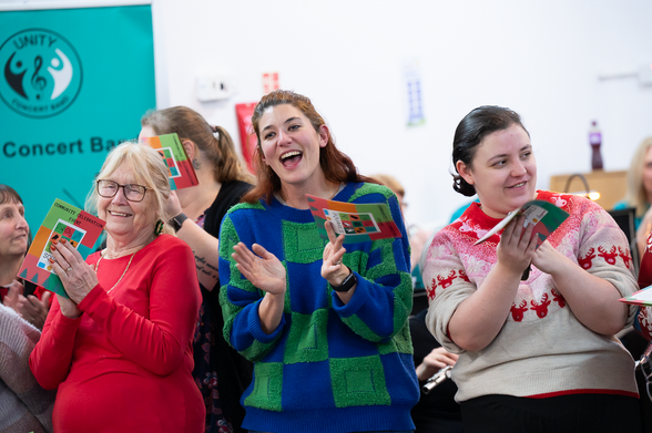 Three smiling individuals stand in the foreground of a musical gathering. One wears a red top and glasses, holding a colorful booklet; another claps joyfully in a blue-green checkered sweater; the third wears a cream sweater with red patterns. A teal banner in the background reads “UNITY Concert Band.”