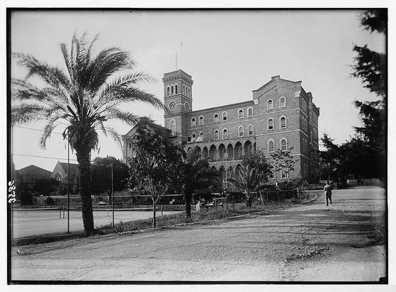The image depicts a historical building, which appears to be College Hall at the American University of Beirut (AUB) during approximately 1920-1933. The structure is an impressive three-story brick edifice with numerous arched windows and decorative masonry details indicative of early 20th-century architecture in Lebanon.
The photograph captures a black-and-white scene, likely taken on a sunny day given the shadows cast by trees and clouds visible through them. In the foreground, there's a barren road or courtyard leading to College Hall, with a few individuals walking leisurely along it. A solitary palm tree stands prominently at the left edge of the frame.
The surrounding environment includes manicured foliage, such as palm trees that add a tropical feel to this urban setting. The image also shows parts of other buildings and infrastructure in the vicinity but remains focused on College Hall itself, showcasing its architectural features like arched doorways and windows, ornate cornices along the roofline, and a clock tower with visible clocks.
The overall atmosphere is serene yet carries historical significance as it captures an institution that likely played vital roles within educational history.