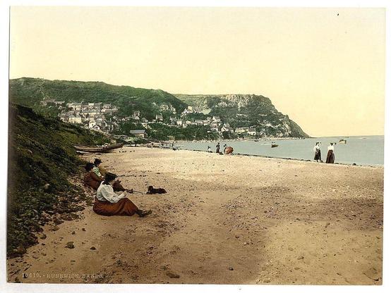 This image depicts a beach scene at Runswick Sands in Yorkshire, England. The photograph appears to be from the late 19th century or early 20th century based on its style and clothing of the people present. On a sandy shore with gentle waves lapping nearby, several individuals are enjoying leisurely activities typical for that era: sitting, walking along the beachfront, and interacting in small groups. In the background rises a steep cliffside dotted with numerous buildings indicative of coastal residences or accommodations catering to tourists.

The photograph is characterized by its sepia tones, which were commonly used during this period due to photographic processes available at that time. The presence of boats suggests recreational boating was popular among visitors. This snapshot offers insight into historical social practices and leisure activities in Britain's seaside resorts around the turn of the century.