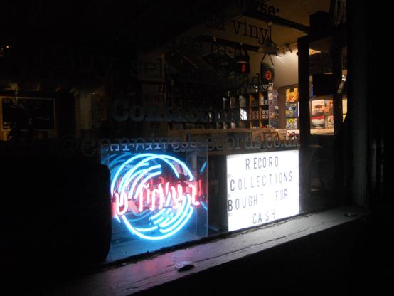 A storefront window at night featuring a neon sign and a light board advertisement. The neon sign displays stylized text, while the light board reads "RECORD COLLECTIONS BOUGHT FOR CASH." The interior shows shelves with vinyl records and promotional material.