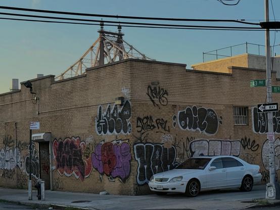 A white car sits idly parked on a sidewalk in front of an industrial building covered in graffiti. Behind it part of the 59th Street Bridge can be seen.