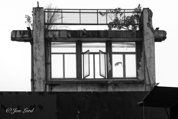 This is an abstract black and white photo in landscape format of a row of three sets of windows left standing on a demolition site. To Kwa Wan, Hong Kong (2017).

In the centre of the image is the row of windows, left standing on the first floor with nothing in view beyond. The windows are set in a thick concrete, weathered and decaying rectangular frame. Each set of the windows is rectangular with two arranged vertically next to each other, with the third set horizontally above the lower two. This pattern is repeated in all three set of windows. The centre two vertical windows are open. Above the windows is a thick concrete lintel with a metal railing stretching across its length: suggesting it might have been a balcony. At each end of the railing is a small amount of wild vegetaion growing. Surrounding this scene is a white background, devoid of any detail, suggesting an overcast sky.