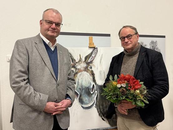 Zwei Männer stehen vor einem Gemälde eines Eselkopfes. Der eine hält einen Blumenstrauß.