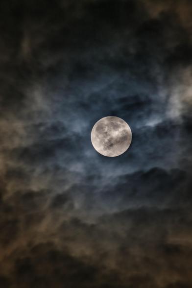 Das Bild zeigt den Vollmond, der leuchtend im Zentrum der Aufnahme steht. Er ist umgeben von dichten, dramatischen Wolken, die ihm einen geheimnisvollen und fast unheimlichen Rahmen geben.
Die Wolken haben dunkle, bläuliche und bräunlich-graue Farbtöne, was dem Bild eine sehr stimmungsvolle, mystische Atmosphäre verleiht. Das Licht des Mondes bricht leicht durch die umgebende Wolkendecke.