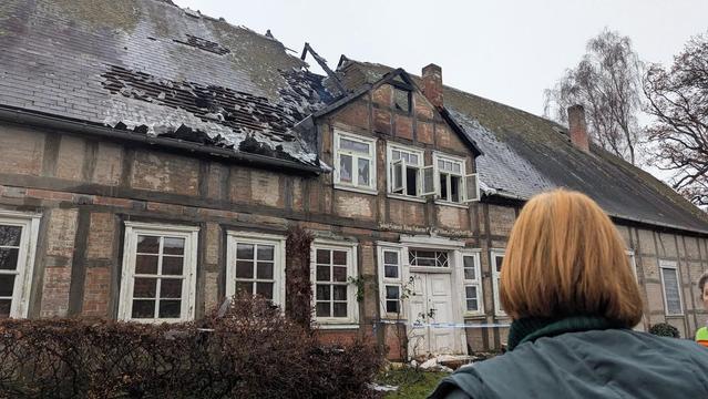 Fachwerkhaus mit beschädigtem Dach nach einem Brand, im Hintergrund Bäume, eine Person steht dem Haus zugewandt.
