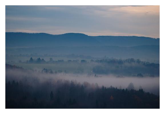 Foto im Querformat. Blick über Wälder, Nebel ist zwischen den Bäumen. Am Horizont erheben sich Berge. Der Himmel ist bewölkt. Die einzigen Farbtupfen im Bild sind einige Bäume mit herbstlich rotem Laub.