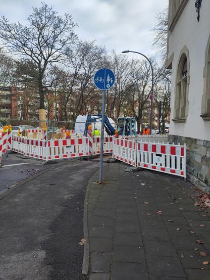 Foto einer Baustellenabsperrung auf einem Fahrrad- und Gehweg, durch die ein ca. ein Meter breiter Durchgang führt, der aber noch immer als getrennter Geh- und Radweg ausgeschildert ist.