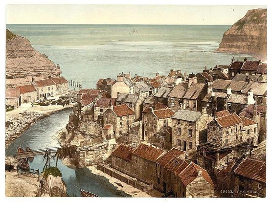 The image depicts an aerial view of the coastal village of Staithes in North Yorkshire, England. This picturesque scene features a collection of traditional stone houses with tiled roofs clustered together along a narrow street that winds its way down to meet the sea.

In the foreground, there is a small river or stream meandering through the town and crossing over it via a wooden bridge supported by metal beams. Alongside this structure stands what appears to be an old watermill with large machinery in operation, possibly for grinding grain or pumping water.

The village extends down towards the shoreline where several boats are docked at the beach. Further out on calm waters, two sailing vessels can be seen near a rocky headland that creates a natural harbor.

In contrast, cliffs rise dramatically from the shore to form part of the landscape's backdrop and provide protection for the town against rough seas. The overall atmosphere conveys a sense of tranquility and timelessness in this historic coastal setting.