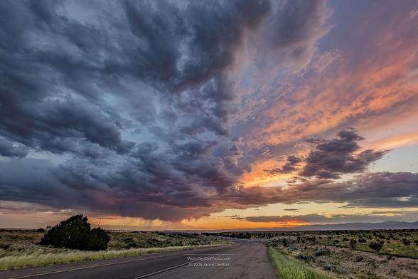 A paved road stretches through a rural landscape under a dramatic sunset sky. Clouds fill the sky, with shades of orange and purple visible. Shrubs and grass line the road.