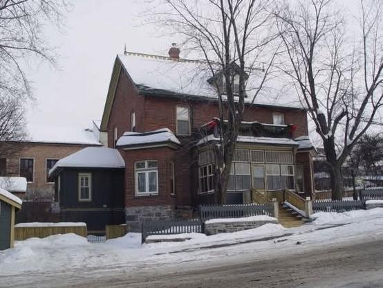 The image displays the old Kendall House in Kenora, Ontario with its roof line covered in snow on a January morning. The old brick building on a stone foundation has carefully painted trim and a closed-in porch at the top of a wide painted wood staircase.