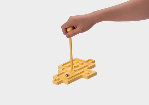 A hand moves a yellow maze toy with a stick, guiding a small ball through its winding paths.