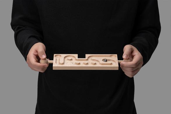 A person holds a wooden maze toy, guiding a metal ball through its winding paths, against a gray background.