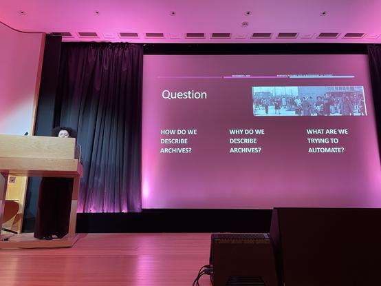 A light-skinned Black woman is just visible behind a podium on a stage. Her slides ask:
How do we describe archives?
Why do we describe archives?
What are we trying to automate?