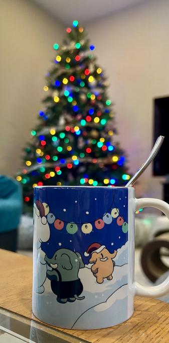 A festive mug featuring Mastodon cartoon characters celebrating the holidays in a snowy landscape, with a colorful Christmas tree, slightly blurred, adorned with lights in the background.