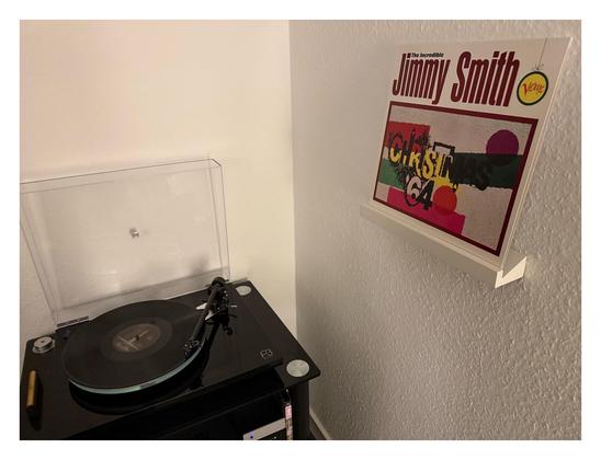 Auf einem schwarzen Plattenspieler läuft eine schwarze Schallplatte. Rechts an der Wand ist ein kleines weißes Regalbrett, darauf steht das Cover. Weiß, oben der Interpret, daneben das Label Logo unten der Titel grafisch stilisiert.