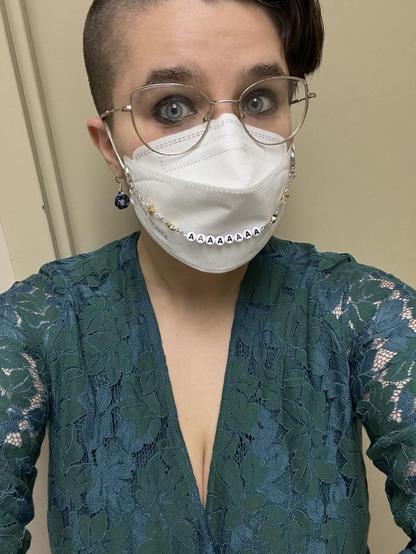 selfie of a young white person with short brown hair and glasses (me). i’m wearing a lacy green dress, earrings in the colors of the bi flag (though the colors are hard to see) and a white mask with a silver and gold mask chain that has letter beads spelling out AAAAAAAA.