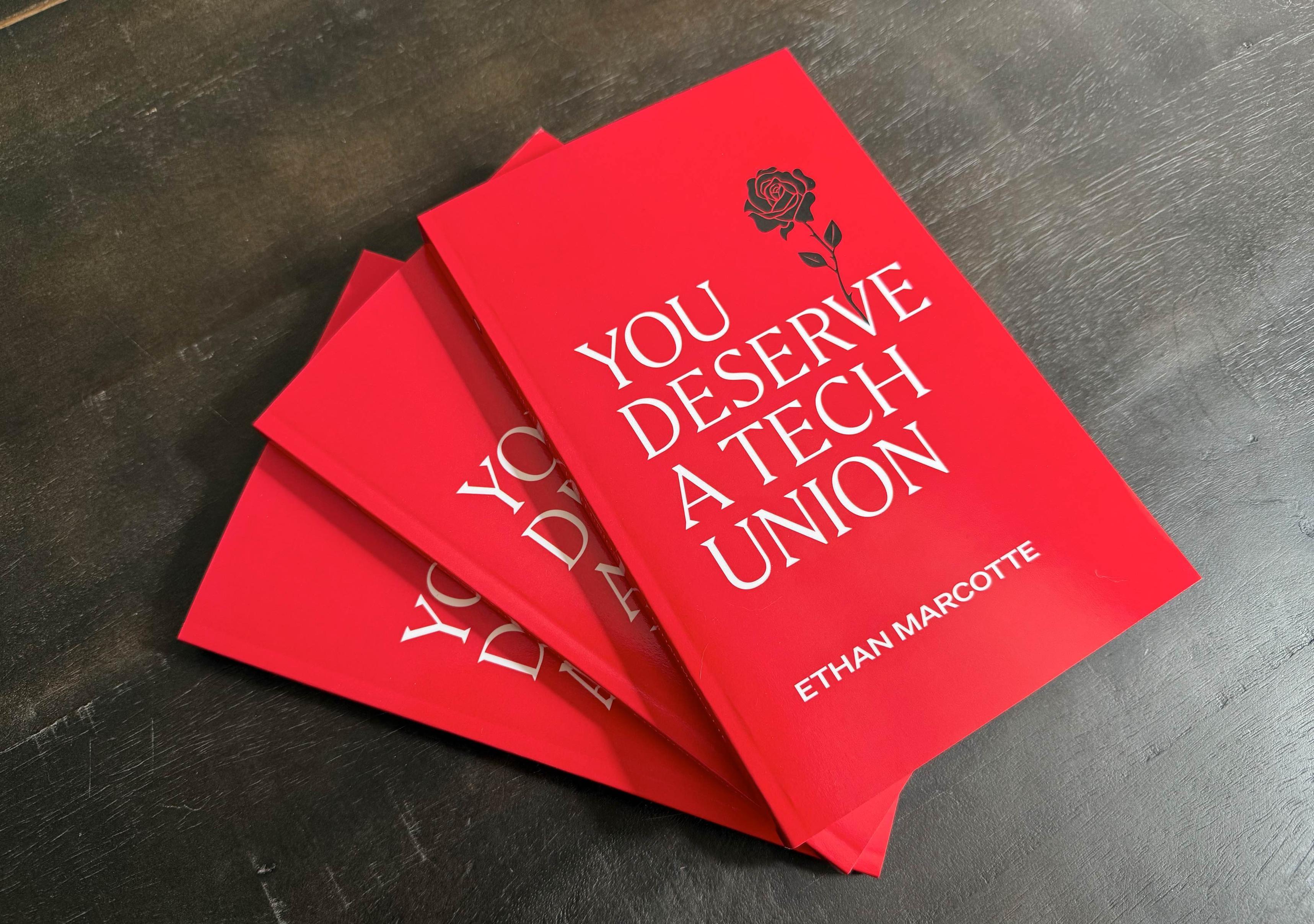 Three bright red copies of the book “You Deserve a Tech Union” are stacked on a gray wooden surface. The stack is flared slightly, so that the corners of the lower books are peeking out from below.