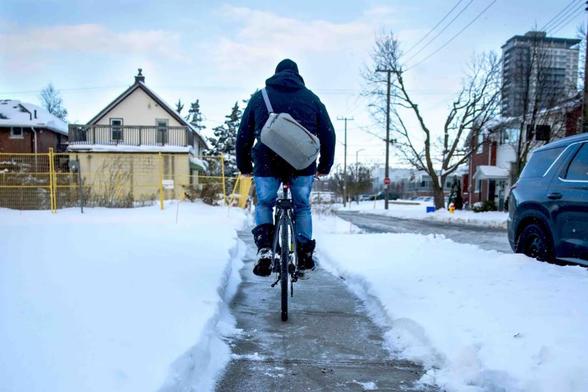 Photo taken outside of a man in a heavy winter coat and hat with a messenger bag cycling down a straight sidewalk away from the camera while flanked by snowbanks.