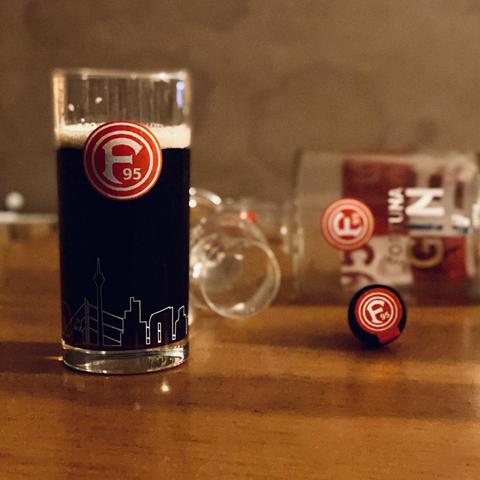 Ein Altbierglas mit Fortuna Düsseldorf Logo, dahinter eine liegende Flasche mit Fortuna Gin, ein liegendes Schnapsglas und ein Korken mit Fortuna Logo