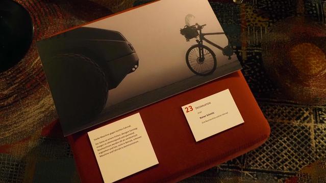 Bild in grautönen, auf dem eine Baumaschine und ein Fahrrad mit geschmücktem Lenkerkorb gegenüberstehen.
Da drunter die Ausstellungsnummer mit dem Titel: Deeskalation
Name des Fotografen