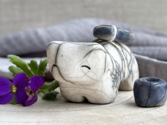 a raku ceramic white bear sculpture with a kind, gentle face. it carries a blue ceramic crescent moon on its back and has a little blue bowl too. it's standing on a piece of pale wood with concentric grain rings, and there's grey muslin cloth and an out-of-focus linen backdrop behind it. there are two small purple flowers with green leaves next to the bear on the left.