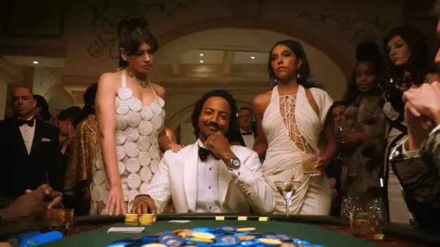 Promotional still from Pluribus Episode 6 showing Diabaté (center) in a white tuxedo seated at a casino poker table with stacks of blue and yellow chips. He's flanked by two women in elegant white evening gowns—one in a geometric circular-patterned dress and one in a draped halter dress. Behind them stand formally dressed attendees in a luxurious casino setting with arched doorways and ornate lighting. The scene depicts wealth, power, and the glamorous aesthetic of the Hive Mind world.