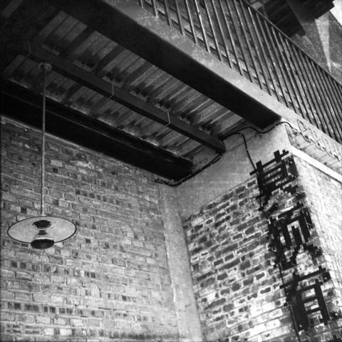 Ilford HP5 Plus 400 (6x6)
English Alt Text: A gritty black and white photo shows an urban scene with a brick wall, an industrial-style hanging light, and a metal ceiling structure with beams. A balcony with vertical railings is visible above. On the right side of the wall, Chinese characters read “Non-existent” adding a personal or rebellious tone. The image blends industrial architecture with expressive street art.
中文替代文字: 这是一张黑白照片,画面呈现都市风格:砖墙、工业风吊灯和带有横梁的金属天花板结构。上方可见带有垂直栏杆的阳台。墙右侧写有中文“莫须有”,传达出个人或叛逆的情绪。整张图融合了工业建筑与富有表现力的街头艺术。