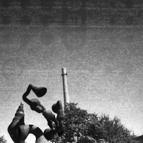 Ilford HP5 Plus 400 (6x6)
English Alt Text: A black and white photograph features a metal abstract sculpture in the foreground, composed of twisted tubular forms resembling horns or funnels. Behind it stands a tall industrial chimney, rising vertically into a grainy sky. Trees surround the sculpture, adding contrast between nature, art, and industry. The sculpture’s flowing shapes juxtapose the rigid geometry of the chimney, creating a visual tension between creativity and functionality.
中文替代文字: 这是一张黑白照片,前景是一座金属抽象雕塑,由扭曲的管状结构组成,形似喇叭或漏斗。背景中是一根高耸的工业烟囱,垂直延伸至颗粒感的天空。雕塑周围有树木,形成自然、艺术与工业之间的对比。雕塑的流动形态与烟囱的刚性结构形成视觉张力,展现创意与实用之间的冲突。