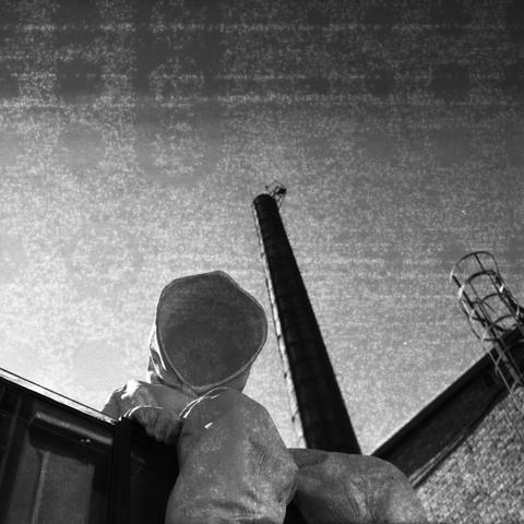 Ilford HP5 Plus 400 (6x6)
English Alt Text: A black and white photo shows a statue of a person wearing a full-body protective suit with a hood, standing on top of a building by the side of a tall industrial chimney. The perspective is from below, emphasizing the towering height of the chimney. The person’s gear suggests a hazardous or contaminated environment. A metal ladder is attached to the building, reinforcing the industrial setting. The dramatic lighting and monochrome tones evoke a sense of caution and seriousness.
中文替代文字: 这是一张黑白照片,画面中一位穿着全身防护服并戴着头罩的人形雕塑坐在一座建筑顶上,旁边是工业烟囱。拍摄角度从下往上,突出了烟囱的高度。所穿装备暗示该环境可能存在危险或污染。建筑上附有金属梯子,增强了工业氛围。黑白色调和光影效果营造出一种警觉和严肃的氛围。