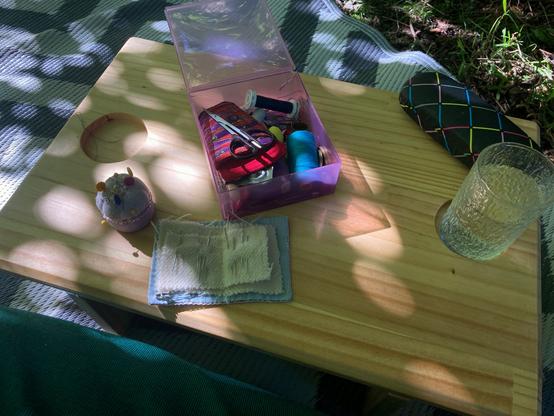 Small wooden table in the shade. A sewing kit with needles, pins, scissor and thread sit on the table. A glass of liquid is at one end, and a glasses case is in the top right corner.