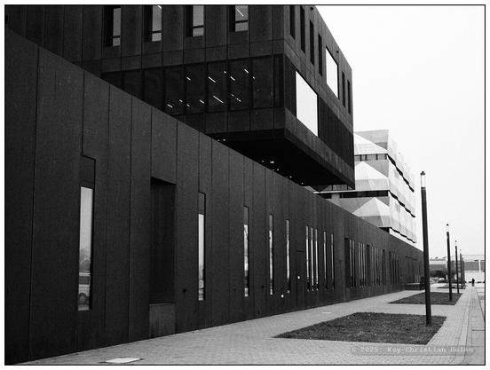 Schwarzweiße, weitwinkligere Aufnahme desselben Gebäudes mit seinen rechteckigen Fenstern und klaren Linien. Rechts neben der Fassade verläuft ein gepflasterter Gehweg mit einzelnen Laternenpfählen. Im Hintergrund sind weitere Gebäude und ein offener Himmel zu sehen.