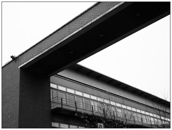 Schwarzweiße Aufnahme eines sachlich-schlichten Bürogebäudes mit langem Fensterband und Brücke aus Backstein im Vordergrund.