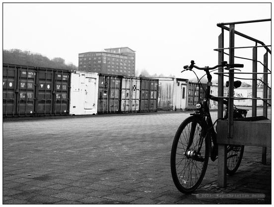 Schwarzweiß-Fotografie eines Fahrrads, das an einem Geländer auf einem gepflasterten Platz lehnt, im Hintergrund in einer langen Reihe aufgestellte Lagercontainer und ein mehrstöckiges Gebäude.