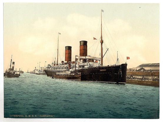 This image depicts a large ocean liner named "Campania," part of the Cunard Steamship Line, docked at Liverpool's port. The ship has two tall red funnels and several smaller masts with flags flying from them. It is surrounded by other ships in what appears to be an active harbor or waterfront area. In the background, there are buildings that may belong to a shipping yard or warehouse facilities associated with maritime activities.

The photograph seems to have been taken during daylight hours under partly cloudy skies. The water surrounding the ship has ripples and waves, suggesting some movement of the sea at this time. This vintage image provides insight into early 20th-century ocean travel, specifically in Britain's port cities like Liverpool.