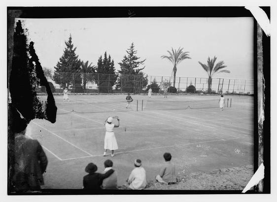 The image is a black and white photograph, possibly an old postcard or print. It depicts a scene of tennis on a court. There are several figures engaged in playing tennis. Two players are at the net, one on each side, while others are spread around the court, some likely waiting for their turn to play. The court is marked with lines indicating boundaries and service boxes.

On the sidelines, there's a group of spectators seated and standing, attentively watching the game. The spectators include both men and women. In the background, there are palm trees swaying gently, suggesting a tropical or Mediterranean location.

The image has a vintage feel to it due to its monochromatic color scheme and possibly the style of clothing worn by the individuals. There is a noticeable tear in the lower right corner of the image, indicating it may have been damaged or aged over time.