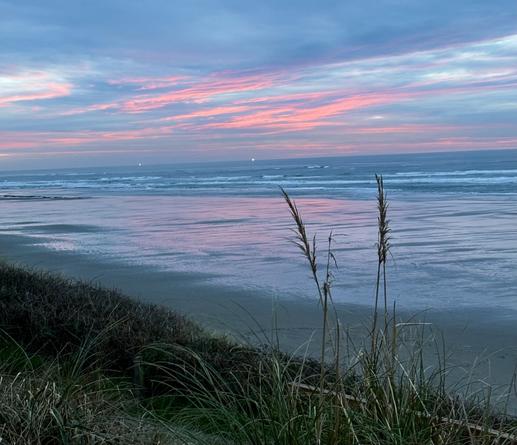 Two distant lighted fishing boats on the horizon as the sun sets. Pink clouds reflect on the wet beach as buts of blue peak out from the clouds. Photo by Robert Emond.